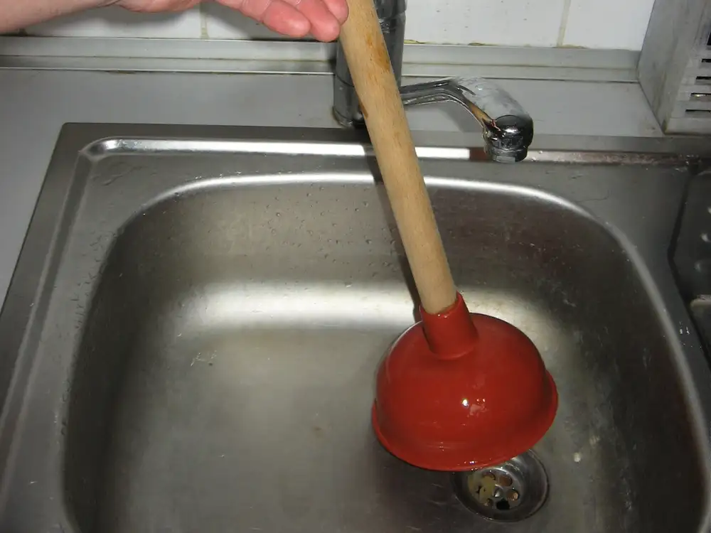 Una mano usa un desatascador de ventosa roja sobre el desagüe de un fregadero de acero inoxidable