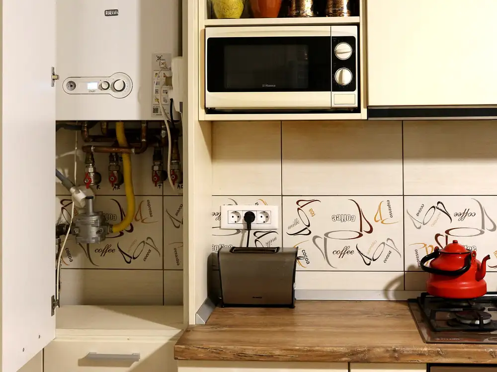 Caldera mural de gas instalada dentro de un mueble alto de cocina, con las tuberías y llaves a la vista, junto a un microondas